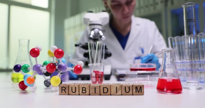 Wooden cubes spell word Rubidium in bright science lab. Female scientist uses microscope and colorful molecule model explaining element behavior