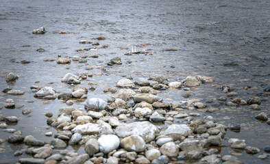 Obraz premium Rocky shoreline with smooth pebbles and stones partially submerged in calm water, creating a natural landscape along a riverbank or lakeshore
