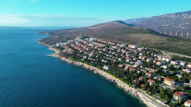 Novi Vinodolski's vibrant marina and rocky Adriatic coastline under a clear winter sky, with a unique cloud formation crowning distant mountains. A picturesque Croatian coastal town.