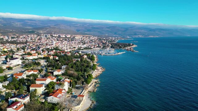 Novi Vinodolski's vibrant marina and rocky Adriatic coastline under a clear winter sky, with a unique cloud formation crowning distant mountains. A picturesque Croatian coastal town.