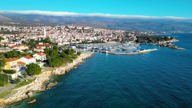 Novi Vinodolski's vibrant marina and rocky Adriatic coastline under a clear winter sky, with a unique cloud formation crowning distant mountains. A picturesque Croatian coastal town.