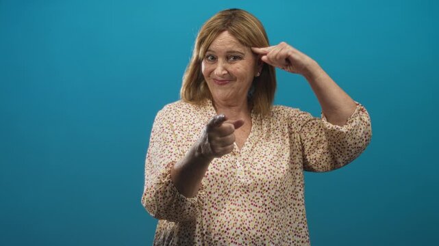 Woman pointing finger at camera and touching temple with other hand, wearing floral blouse against blue studio backdrop; confident wit.