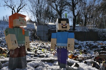 Naklejka premium Minecraft figures of Steve and Alex standing in winter garden with sun behind their backs, first snow on the ground. Older steel water barrrel and fruit trees visible in background. Clear blue skies. 