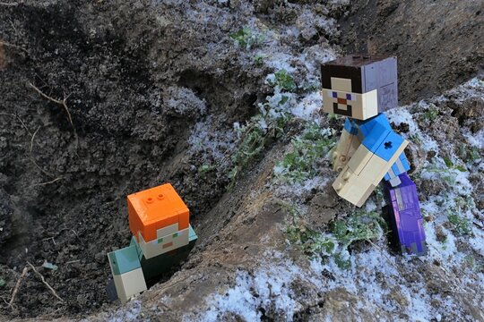 Minecraft action figure of main character Steve watching main character Alex trying to climb out of dug out garden pit, partially frozen soil visible around. 