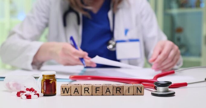 Wooden cubes spell word Warfarin on tidy hospital desk. Female doctor writes detailed prescription near stethoscope and red scattered capsules