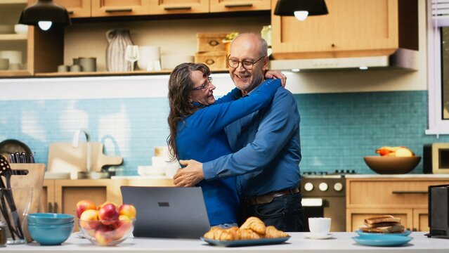 Smiling aged husband and wife sharing a romantic dance in the kitchen, waltzing together on a passionate love song and flirting with each other. Holding hands and slow dancing, swaying. Camera B.
