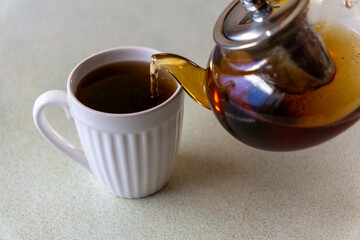 Close-up of hot tea being poured from a transparent glass teapot into a white ceramic mug on a clean, minimalist kitchen surface. Warm tones, natural light, and a calm atmosphere convey comfort, relax