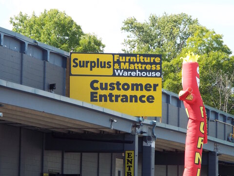 Ottawa, ON, Canada-July 11,2025: A yellow "Surplus Furniture & Mattress Warehouse" sign marks the customer entrance next to a red inflatable tube man on  St. Laurent Blvd.