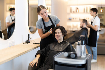 Young woman barbershop employee stands next to female client, discussing details of haircut. Stylist and client agree on haircut, master attentively listens to wishes of visitor.