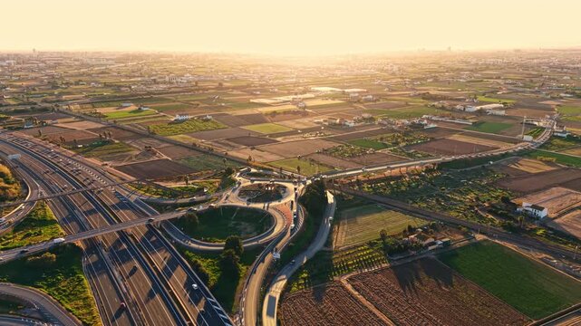 The scene shows a wide view of an agricultural area during sunset. There are fields divided into sections and a highway with cars. The sky has warm colors as the sun sets.