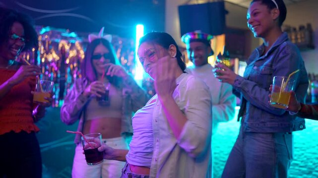 Happy young woman with sunglasses holding a drink while dancing with a diverse group of friends in a crowded nightclub. Enjoying the vibrant music and energetic atmosphere under colorful neon lights