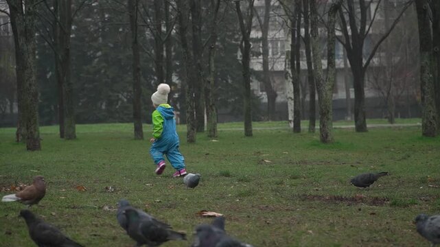 A child in a blue-green jumpsuit, holding a shovel and a bucket, runs cheerfully toward a flock of pigeons that his mother is feeding to scare them away. A rainy autumn day.
