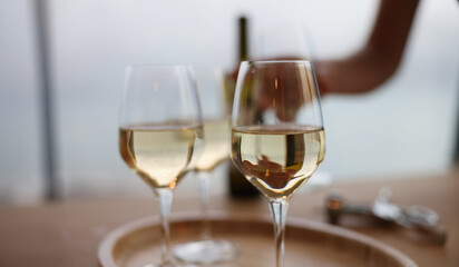 Naklejka premium Charming white wine glasses sit on a wooden tray, shimmering in the soft light. A gentle hand reaches for the bottle, enhancing the tranquil atmosphere of this midday gathering by the water.