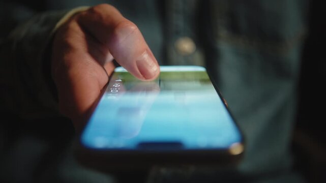 Macro shot of male hand in blue denim shirt scrolling social media feed on smartphone at night. Swiping finger, motion blur on phone screen. Evening online browsing, digital content consumption