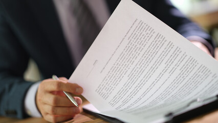 A diligent individual in a suit examines detailed documents with a pen in hand. Natural light filters through, highlighting their concentration and the organized workspace.