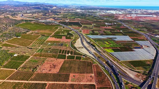 An aerial view shows large fields with rows of crops and open land near a highway. In the distance, mountains and a coastal area are visible under clear skies.