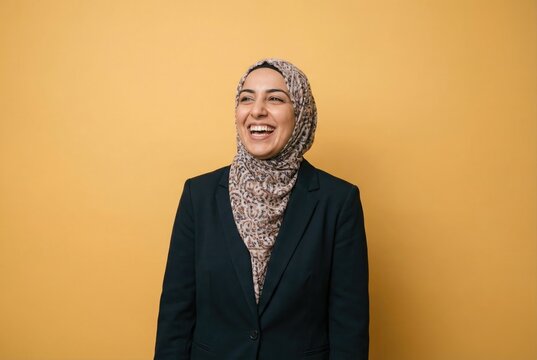 A cheerful Muslim businesswoman in a hijab and suit laughing heartily on a yellow background.
