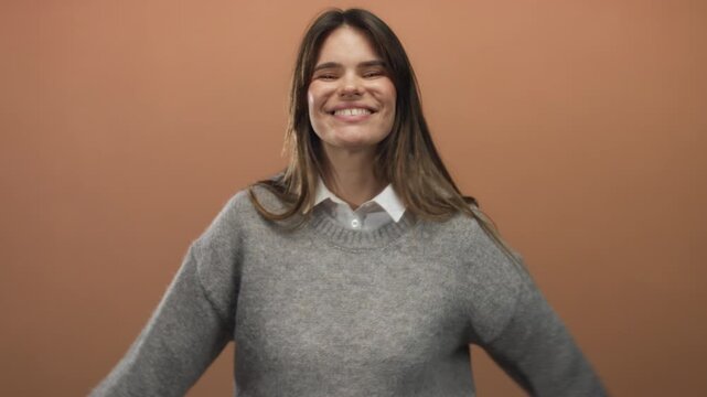 Woman smiling in gray sweater against warm brown background sending a cheerful airborne kiss showcasing young hispanic joyful expression creating a friendly isolated atmosphere.