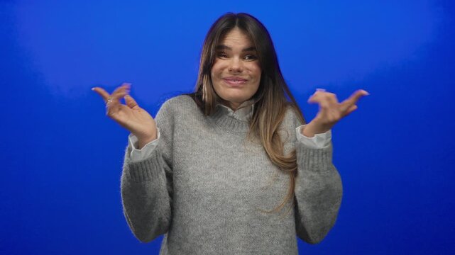 Hispanic woman in grey sweater gesturing in confusion against a solid blue background, showcasing emotions and expressions in an isolated setting.