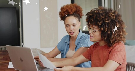 Reviewing papers, two women wearing denim shirt and rust top, glasses, using silver laptop at home