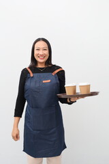 Happy Asian female barista wearing blue apron, standing holding tray with coffee paper cups, smiling looking at camera, isolated on white background.