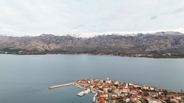 Wide drone panorama with a slow turn over a small boat marina in Vinjerac, showing fishing boats, the town, calm sea and snowy Velebit mountains on an overcast winter day. Zadar region, Croatia