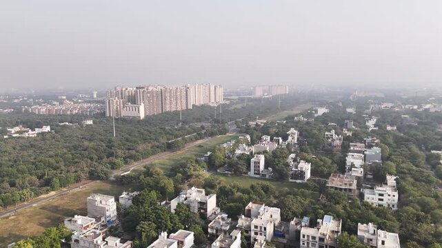 Wide drone shot showcasing a green urban corridor flanked by housing societies and open land, highlighting planned development, road connectivity, and morning haze over the expanding cityscape.