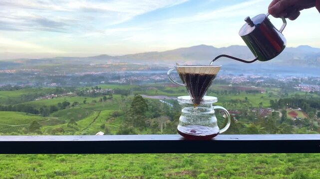 Hand pouring water from gooseneck kettle into glass coffee dripper overlooking scenic misty mountain valley