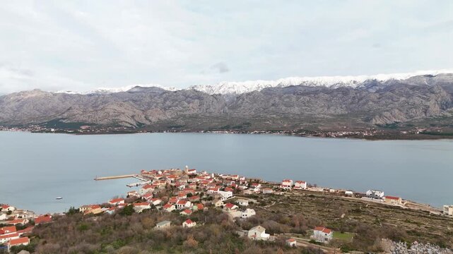 Drone panorama of Vinjerac on an overcast winter day, showing red rooftops by a calm Adriatic Sea with snow-covered Velebit mountain peaks in the background. Zadar region, Croatia