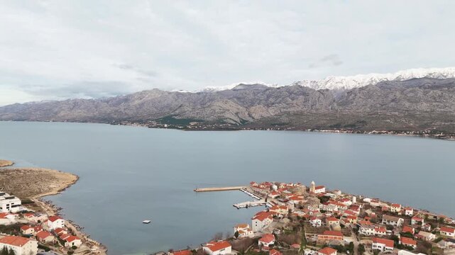 Drone footage flying directly toward a pier and sheltered cove in Vinjerac on an overcast winter day, with a church, red rooftops and snow-covered Velebit mountains in view. Croatia