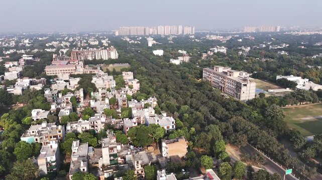 High-angle drone shot showing low-rise housing clusters and vacant land on the outskirts of a dense city, framed by greenery and smoggy skies typical of Delhi NCR.
