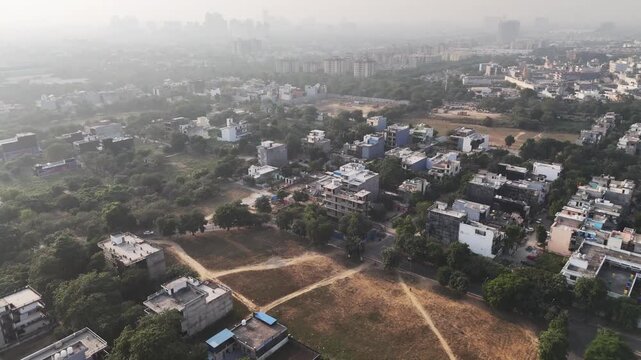 Aerial view of a quiet residential pocket in Delhi NCR, with scattered homes, open plots, and tree cover fading into distant urban haze&mdash;capturing suburban growth amid visible air pollution.
