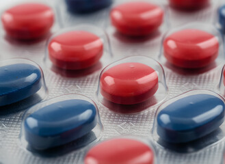 A close-up macro photograph featuring vibrant red and blue oval pills encased in a transparent plastic blister pack with silver foil backing. This image represents the pharmaceutical industry.