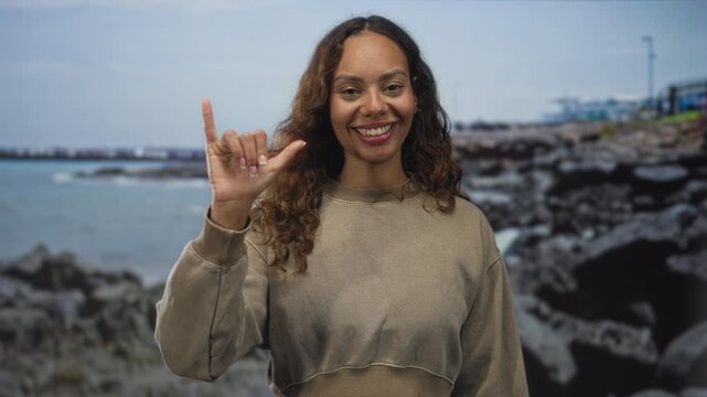 Woman smiling with hand making l sign in a studio seaside set showing rocky coast and water; friendly greeting.