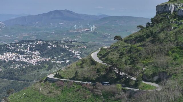 AERIAL: Series of switchbacks cuts across a green mountainside, overlooking a valley town and a wind farm in the distance. A bus and car trace the serpentine route beneath a sunny Mediterranean sky.