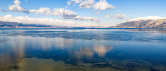 Fototapeta premium Panoramic aerial view of a serene lake with crystal clear water reflecting the blue sky and distant snow covered mountains during winter