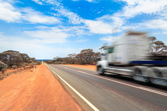 Long straight road through the Australian outback with motion blurred road train passing under blue sky, showing remote rural landscape in Australia.