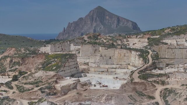 AERIAL: Marble quarries carved into Sicilian hills, overlooking the blue Mediterranean Sea. White rock faces, winding service roads, and scattered buildings contrast with green scrub and coastline.