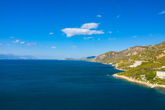 Vivid view of a deep blue sea stretching alongside a rugged, forested coastline, with sunlit hills and scattered buildings under a bright, cloud-dotted sky. The scene radiates clarity, freshness, and