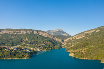 Fototapeta premium Aerial view of a lakeside village nestled among green hills and dramatic cliffs along the turquoise waters of Lac de Castillon, with a prominent mountain peak in the distance and clear summer skies