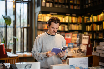 Fototapeta premium Focused man booklover reading popular book standing in middle of local bookstore, surrounded by various publications on display. Thoughtful male choosing book in bookshop, looking through bestseller.