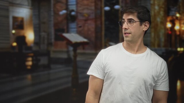 Man points finger toward lectern in building wearing glasses and white tshirt; sincerity reflection.