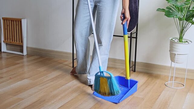Person in jeans sweeping wooden floor with blue broom and dustpan
