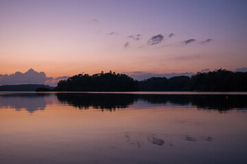 Fototapeta premium Gentle evening light casts soft purple and peach tones across the tranquil surface of Lac des Settons, reflecting the dark outline of distant trees and a serene sky.