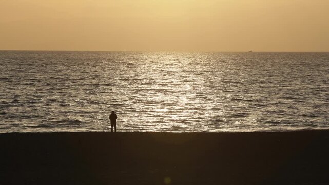 K Wide shot of people walking along the beach at sunset in Chigasaki, Japan. Beautiful golden hour ocean scenery with silhouettes of people enjoying a stroll.