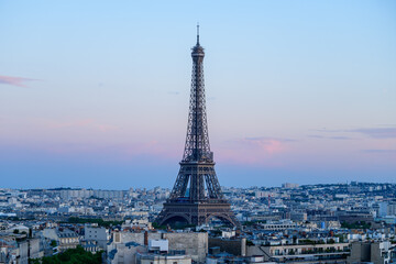Obraz premium Iconic Eiffel Tower stands tall above the Paris skyline, framed by soft pastel hues and scattered clouds at dusk, with city rooftops stretching into the distance.