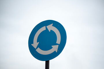 Blue roundabout road sign with white arrows indicating circular traffic flow against a white sky.