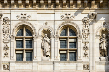 Fototapeta premium Detailed stone statues stand in niches between arched windows on the richly decorated facade of Hotel de Ville in Paris. Sunlight highlights the intricate carvings, floral motifs, and geometric window