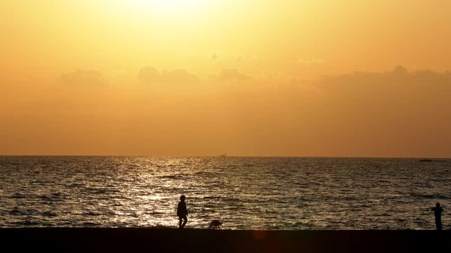 4K Wide shot of people walking along the beach at sunset in Chigasaki, Japan. Beautiful golden hour ocean scenery with silhouettes of people enjoying a stroll.