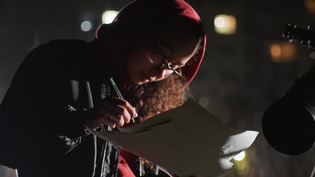 hooded stargazer rooftop night studies star chart amateur astronomer referencing paper map under telescope lamp, city lights bokeh, concentrated expression, slow hands tracing constellation sketches,